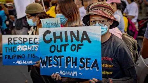 Getty Images A proester in Manila, wearing a hat, eyeglasses and face mask carrying a blue sign with the words China out of West Philippine Sea in bold black font