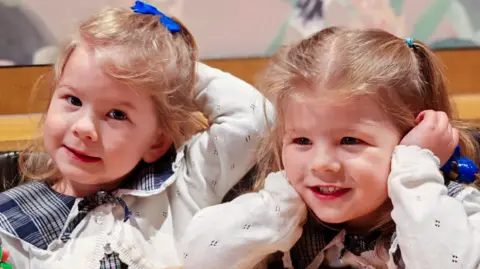 Brioney Garrett Nancy and Margo are photographed close up - their faces and blond hair in bunches are visible, and they are smiling. They are wearing the same clothes - a blue blouse and white cardigan.