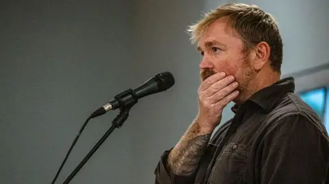 Getty Images Graham Platner seen on stage at a microphone. his hand is over his mouth 