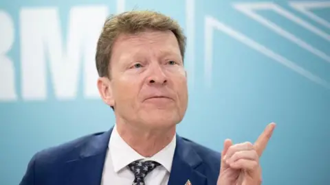 PA News Richard Tice, in blue jacket, white shirt, and black and white tie, points with his finger while talking to reporters     