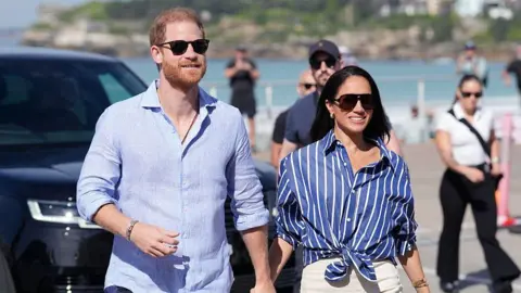 Getty Images Prince Harry, Duke of Sussex, and his wife Meghan, the Duchess of Sussex walking hand in hand, wearing blue shirts and sunglasses.