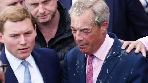 Getty Images Reform UK party leader Nigel Farage reacts after a woman threw a drink over him. On his left is a second man, dressed in a suit with a white shirt and blue tie.