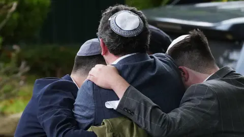 Christopher Furlong/Getty Images Three men wearing Jewish skullcaps - known as a kippahs - embrace in grief on the street after the attack