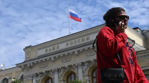 Anadolu via Getty Images A woman wearing a red jumper and sunglasses, as well as a little bag on her right shoulder, talks on the phone in front of an exterior view of the Central Bank of the Russian Federation building on top of which the Russian flag can be seen