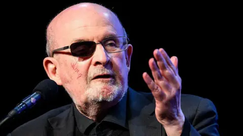 Getty Images Rushdie speaking in March 2026 into a microphone. He has a darkened lens over his wounded eye