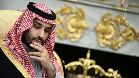 AFP via Getty Images He is rubbing his chin, thinking. He is wearing traditional Arab clothing, with the red and white head dress. He is wearing a black robe with gold edges.