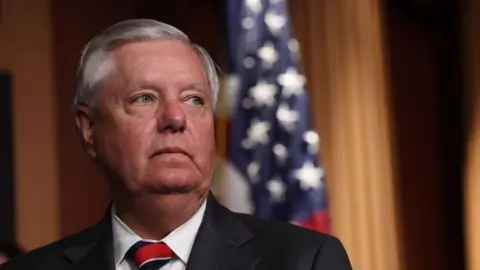 Getty Images Lindsey Graham, a man with short, grey hair, wears a black suit with a white shirt and a red and black tie. He is looking to the right. The background behind him is blurred, with a hanging American flag.