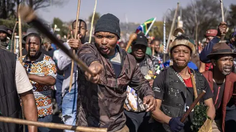 EMMANUEL CROSET / AFP via Getty Images Protesters marching, a man at the front is pointing a stick at the camera.