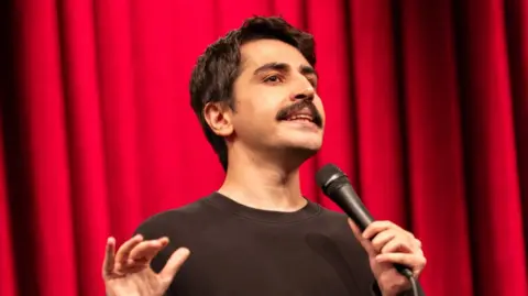 Instagram/@denizgoktasx A head-shot posted by a Turkish stand-up comic showing him holding a microphone and standing in front of a red curtain