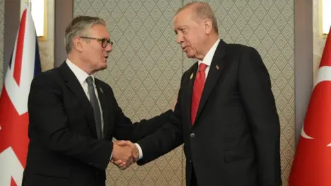 PA Sir Keir and Erdogan, both wearing dark suits, shake hands. Behind them are the Union Jack and Turkish national flag.