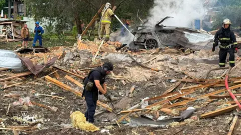 Ukrainian President Zelensky Emergency services clear through the rubble following a Russian missile strike just outside of Kyiv. Firefighters can be seen extinguishing a car fire in the background