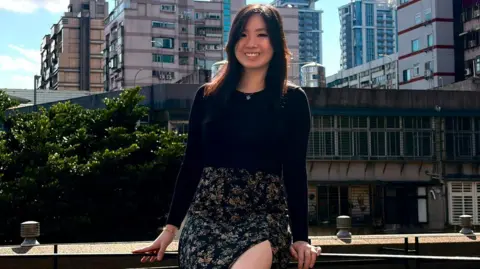 Michelle Huynh Young investor Michelle Huynh posing for a picture while sitting on a fence in front of a blue sky and a housing estate.
