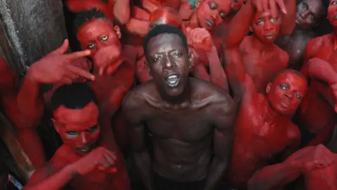 Artists Equity A topless Empereur stands in the centre of a group of young men, who are painted in red.