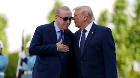 Reuters Donald Trump, dressed in a navy suit and tie, talks into the ear of Turkish President Recep Tayyip Erdogan who is also wearing a blue suit and tie, as well as sunglasses.