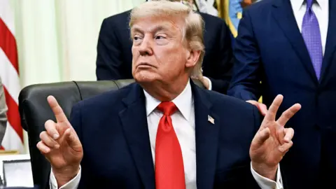 Getty Images US President Donald Trump pointing fingers in the Oval Office of the White House, with an American flag and suited men behind him.