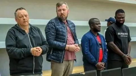 Serviço de Investigação Criminal de Angola Four handcuffed men standing in front of a row of confiscated laptops. Lev Lakshtanov, Igor Ratchin, Amor Carlos Tomé and Francisco Oliveira (left to right)