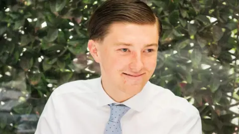 Family photo A young man smiling wearing a white shirt and a blue tie. He is sitting in front of a green bush. He has brown hair.