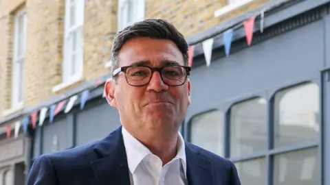 Reuters Andy Burnham looks directly forward and smiles with his mouth closed. He wears tortoise shell glasses, a navy suit jacket is navy and a white shirt. He stands in a street, behind him is a blue shop, with red, blue and white bunting hung above it.
