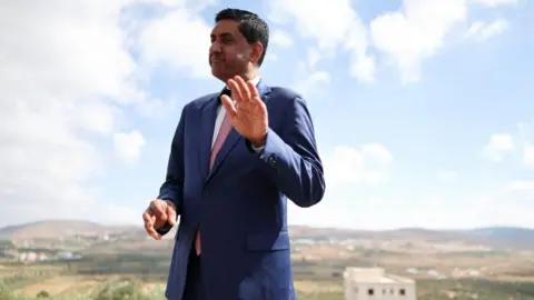Reuters Khanna, who is dressed in a blue suit, white shirt and pink tie, raises his hand as he is photographed outdoors with a rural town landscape behind him
