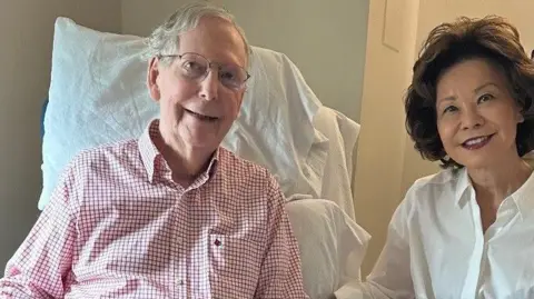 Office of Mitch McConnell Mitch McConnell sits looking ahead in red checkered shirt, next to his wife who is wearing a white shirt