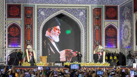 Getty Images A crowd holds their phones up to take pictures at the funeral of Ayatollah Ali Khamenei, a picture of whom appears above a stage. 