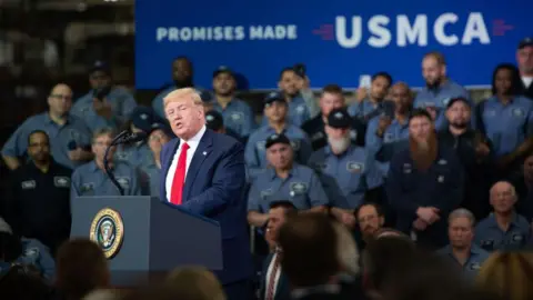 Getty Images US President Donald Trump stands and speaks at a podium in front of auto workers with a "Promises Made USMCA" banner in the top right corner. 