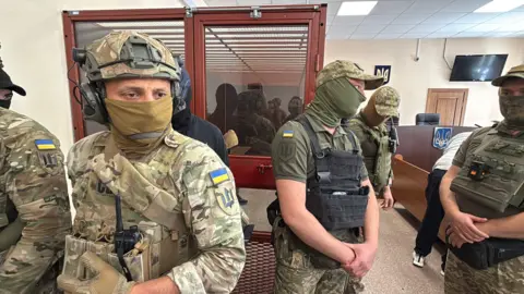BBC Men in military fatigues stand in a courtroom with a person sitting in a glass room