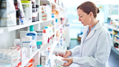 Getty Images Pharmacist standing in front of shelves in pharmacy, holding medicine boxes in hands.