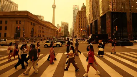 Cole Burston/ AFP/Getty Images People walk on a crossing in a road downtown Toronto. There are buildings in the background and sky is orange from smoke.