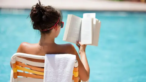 Getty Images Young woman sitting in a chair with a towel draped over the back, reading at the pool in the summer