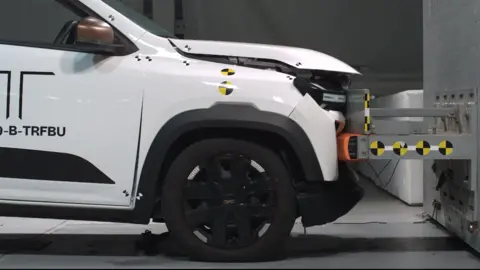 BBC A white electric car crashing into a wall at a low speed in a crash test lab. The front of the car has bent in a bit and the bonnet has lifted up, but otherwise the damage looks minimal.