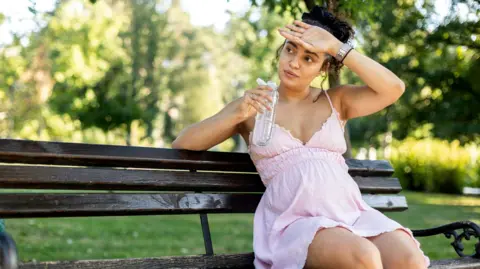 GoodLifeStudio Pregnant woman sitting on a park bench in a light pink dress, holding a bottle of water.  