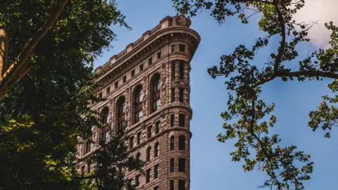 Courtesy of The Flatiron Building A view of the top section of the Flatiron Building, with trees surrounding it, showing its intricate Beaux-Arts detailing