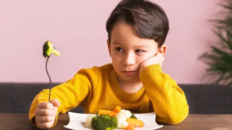Getty Images Young boy in yellow jumper sits at a table with a plate of broccoli, carrots and cauliflower in front of him. He is holding a fork with broccoli on it and looks unhappy. 

