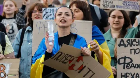 EPA/Shutterstock Ukrainians, including a young woman draped in a Ukrainian blue-and-yellow flag, hold placards reading 'Bring him back" during a protest against the dismissal of Defence Minister Mykhailo Fedorov in downtown Kyiv