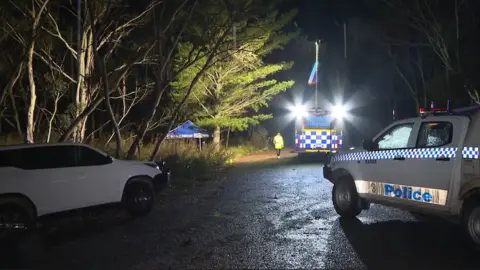 ABC News A night scene on a country road with several police cars and a small blue tent roof behind some trees.