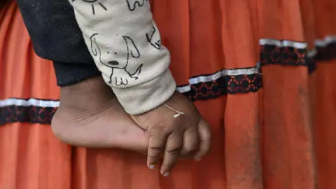 AFP via Getty Images A close-up of a small child’s bare foot and hand is shown against a backdrop of their mother's skirt - a traditional indigenous outfit. It appears the child is being held by their mother. The child is wearing a light-coloured sleeve printed with a simple cartoon dog, and the hand hangs loosely beside the foot.