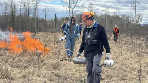 Supplied by Sonja Leverkus A man in an orange hard hat and black workwear carries gas cannister as a team behind him works on a controlled burn. There are four men behind him and they was through dry brush. There is a small fires to the side.
