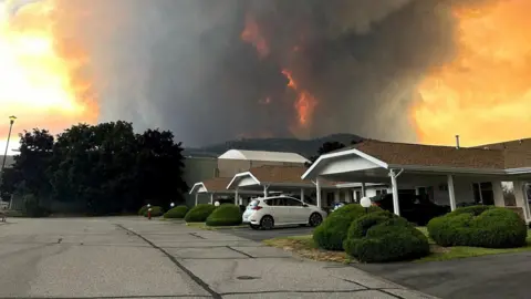 Terry Fries/Handout via Reuters An image showing grey and orange smoke rising from behind a row of homes on a residential street in Summerland, British Columbia. 