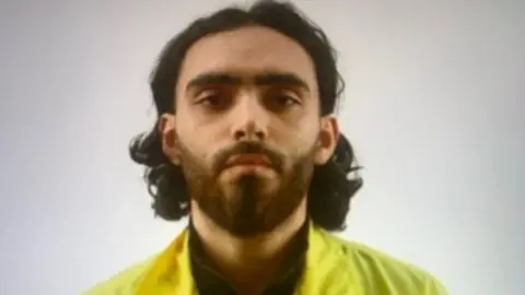 Iraq National Intelligence Service  A man with long dark hair and a short beard in a mugshot-style photo