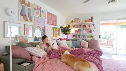 @devonandwillo A woman with her phone sitting on a sofa with multicoloured pillows and covered in a pink blanket next to a dog.