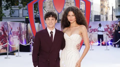 PA Media Tom Holland and Zendaya on the red carpet at the London premiere for Spider-Man: Brand New Day. Holland is wearing a burgundy suit and tie. Zendaya is wearing a white shoulderless dress. 