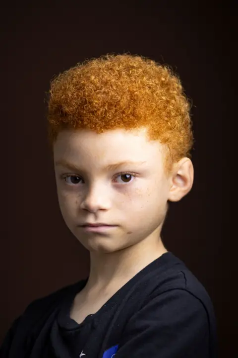 Kieran Dodds Close-up studio portrait of a child with short, tightly curled red hair and freckles, wearing a dark T-shirt. The child faces the camera with the head turned slightly to one side against a dark brown background. Soft, warm lighting highlights the hair texture and facial features.