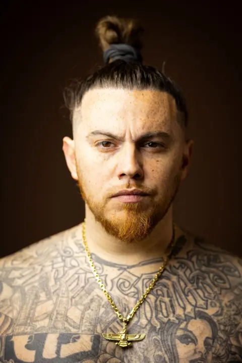 Kieran Dodds Close-up studio portrait of a person with hair tied in a top knot, freckles, a short beard, and extensive black-and-grey tattoos across the chest and shoulders. The person wears a gold chain necklace with a winged pendant and faces the camera against a dark brown blurred background.