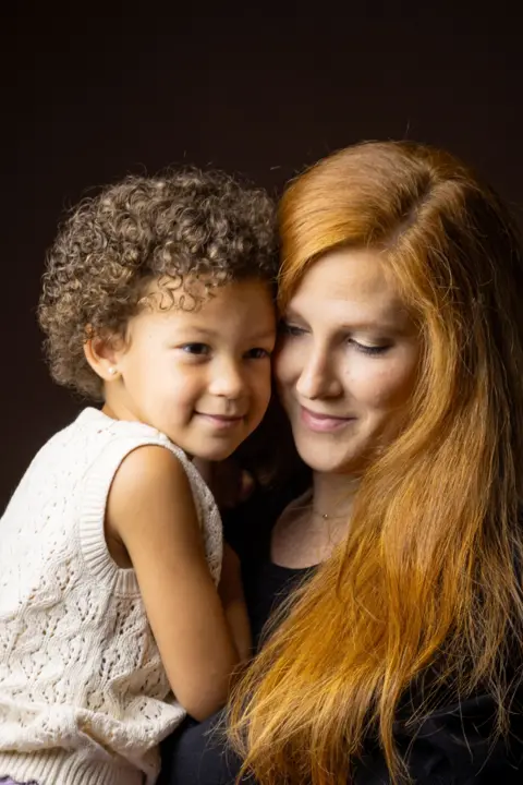 Kieran Dodds Close-up studio portrait of an adult with long red hair holding a young child with short curly hair. The child wears a sleeveless cream knit top, and both are positioned closely together against a dark brown background. Warm lighting highlights the hair texture and clothing details, creating an intimate family portrait.