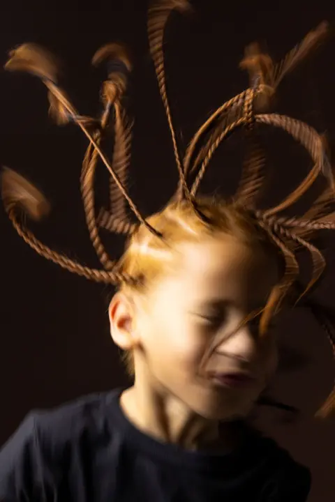 Kieran Dodds Artistic close-up portrait of a child with multiple braided strands of hair extended outward in motion, creating a dynamic blurred effect. The child wears a dark top and is photographed against a dark brown background with warm studio lighting that highlights the movement and texture of the braids.