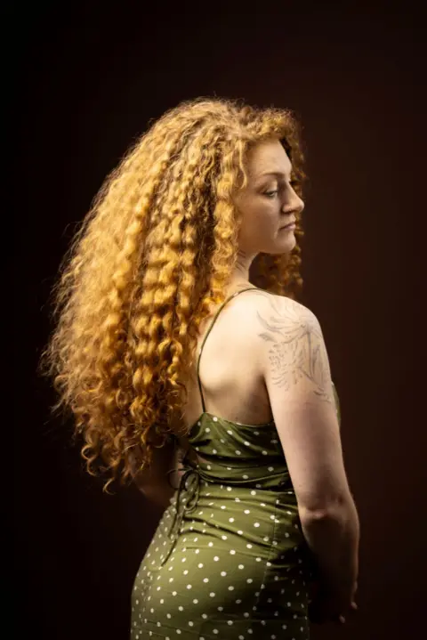 Kieran Dodds Studio portrait of a person with long, voluminous curly hair photographed from the back and side, turning slightly toward the camera. The person wears a green sleeveless dress with white polka dots and thin shoulder straps, featuring a lace-up back detail. A floral tattoo is visible on the upper arm. Warm lighting highlights the texture of the hair against a dark brown background.