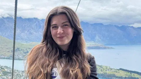 Elena Elena is sitting in a cable car, with a view of a lake and mountains behind her. She is wearing a long top with the sleeves rolled up and has a cream handbag across her body. She has long brown hair and is smiling at the camera, with her hands crossed on her lap.