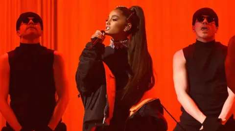 Getty Images Ariana Grande singing on stage with two male dancers standing behind her 