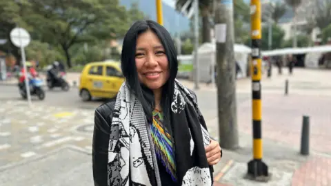 José Carlos Cueto / BBC Mundo A woman with long dark hair stands at a street intersection with trees, vehicles, and pedestrians visible in the background. She wears a black jacket, a large black-and-white patterned scarf, and a brightly coloured beaded garment. She smiles directly at the camera.